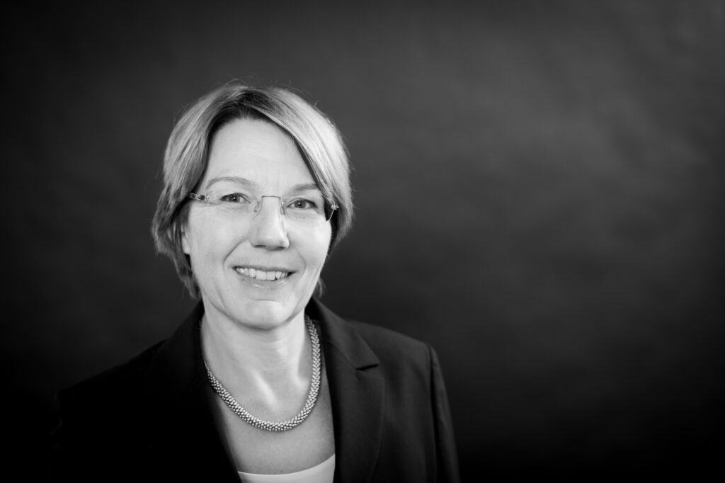 - Porträt einer lächelnden Frau mit kurzen Haaren, Brille und schwarzem Blazer, in Schwarz-Weiß aufgenommen.