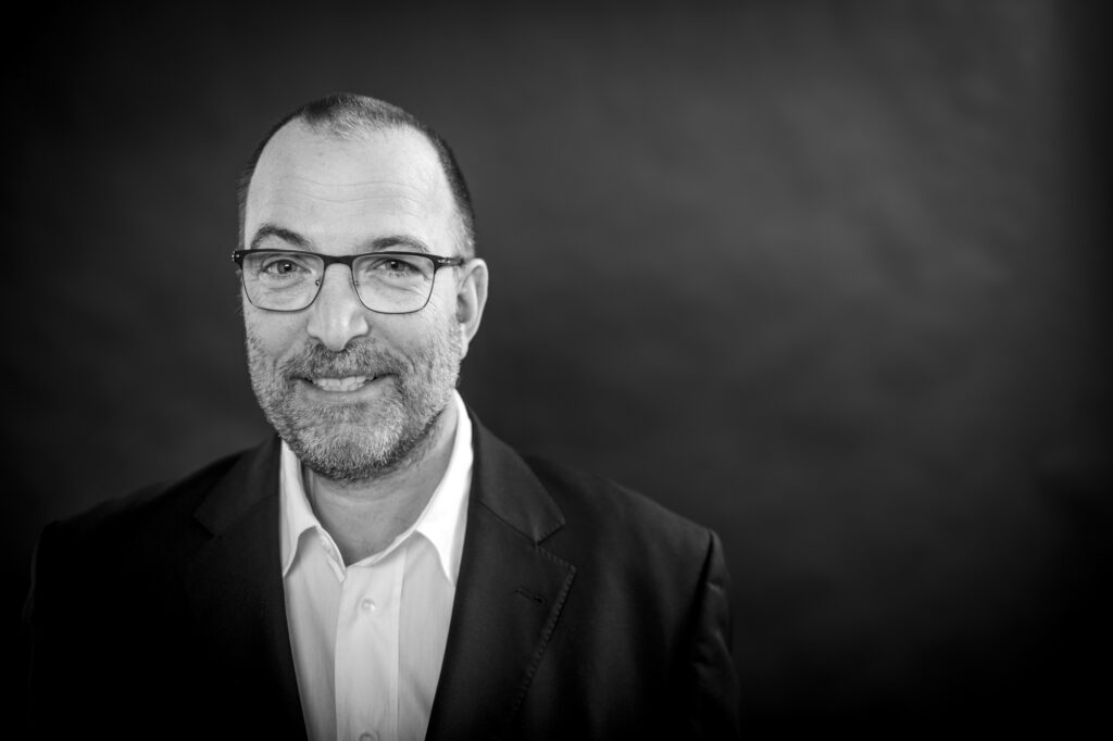 Hochwertiges Portrait eines Mannes mit Brille, professionell aufgenommen in Schwarz-Weiß, vor dunklem Hintergrund.