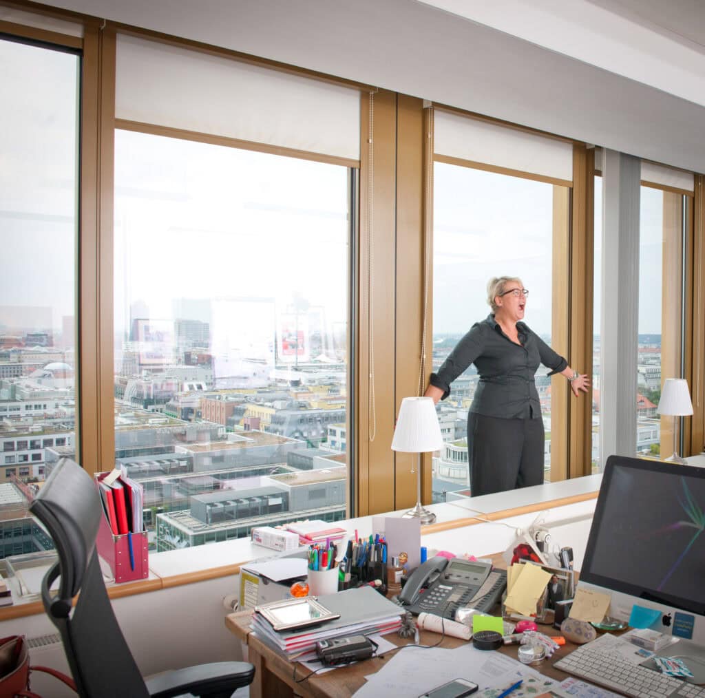 Moderne Büroaufnahme mit großer Fensterfront und Sicht über die Stadt, Frau im Business-Outfit zeigt emotionale Überraschung oder Freude. Arbeitsbereich ist chaotisch mit Akten, Büromaterialien und Technik.