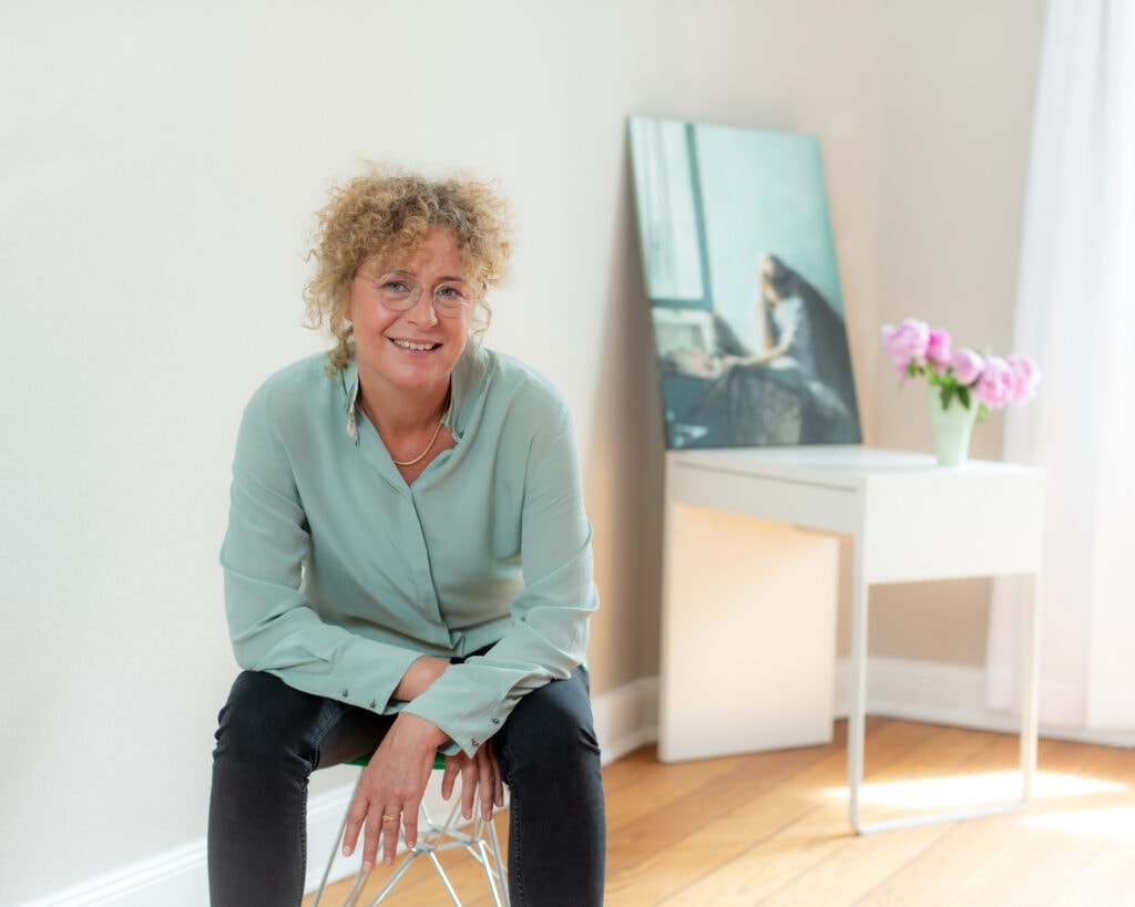 Attraktives Porträt einer Frau mit lockigem Haar und Brille, sitzend in einem hellen Raum, professionell fotografiert von Frank Siemers.