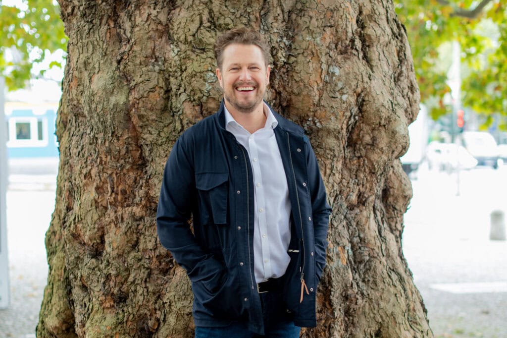 Bild eines lachenden Mannes in einem blauen Jacke vor einem Baum, Porträtaufnahme im Freien.