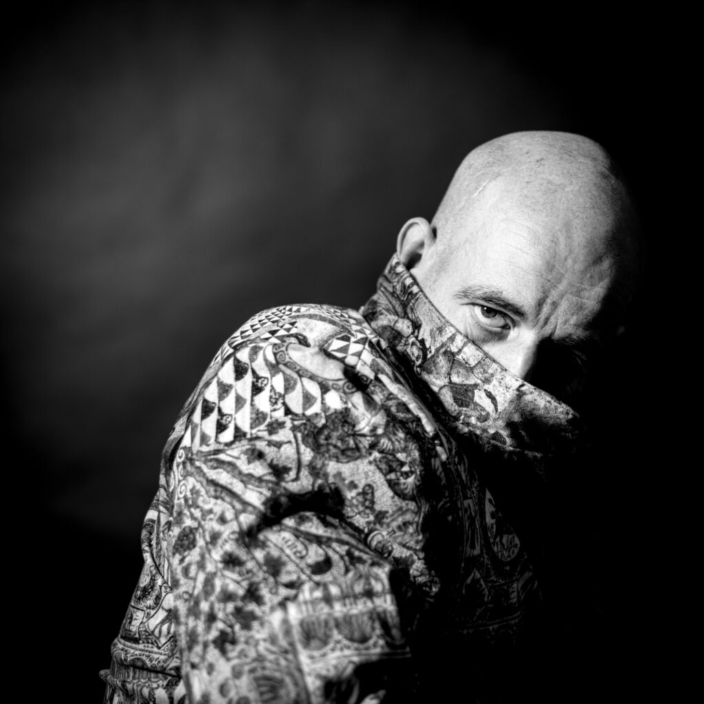 Bilder von Frank Siemers, Hamburg, schwarzer Hintergrund, portraitierter Mann mit intensivem Blick, Tattoos auf seinem Arm, in Schwarz-Weiß.