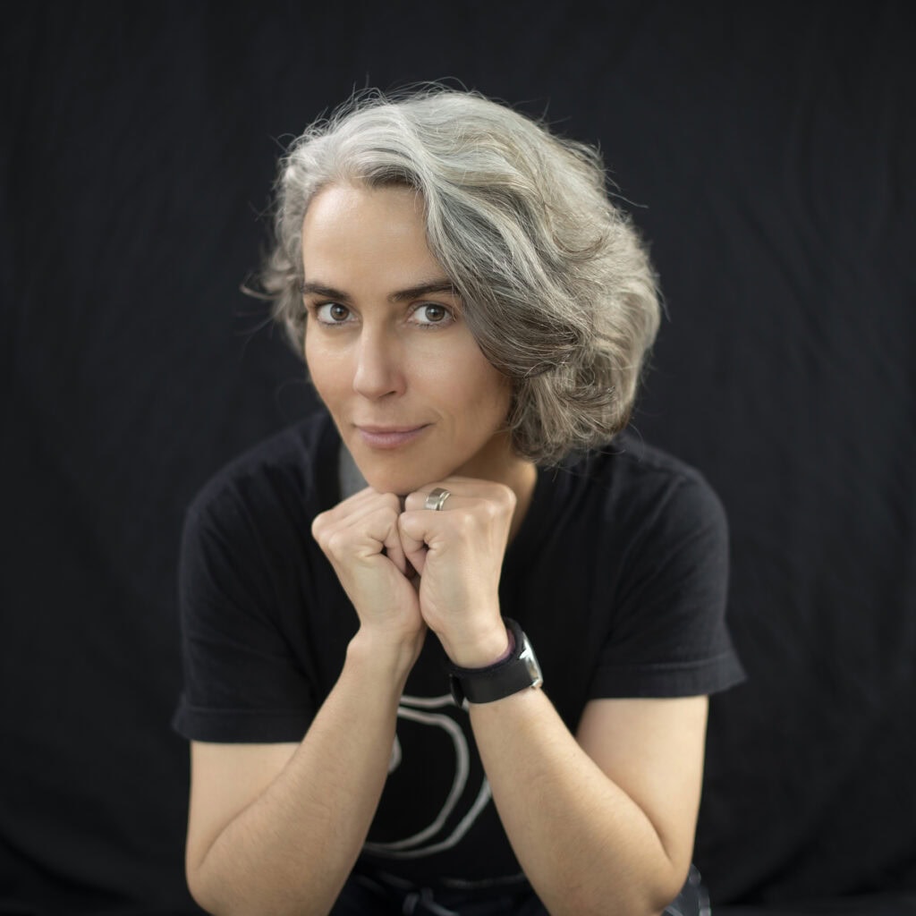 Freundliche Frau mit grauem Lockenkopf und schwarzem T-Shirt, die stolz in die Kamera lächelt, vor schwarzem Hintergrund.