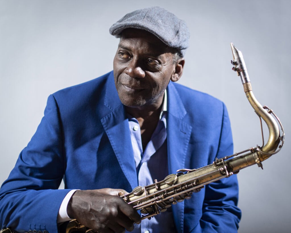 Alter Text: Porträt eines schwarzen Jazzsaxophonisten in blauer Jacke und Mütze, der in die Kamera schaut, mit Saxophon in der Hand, vor hellem Hintergrund.