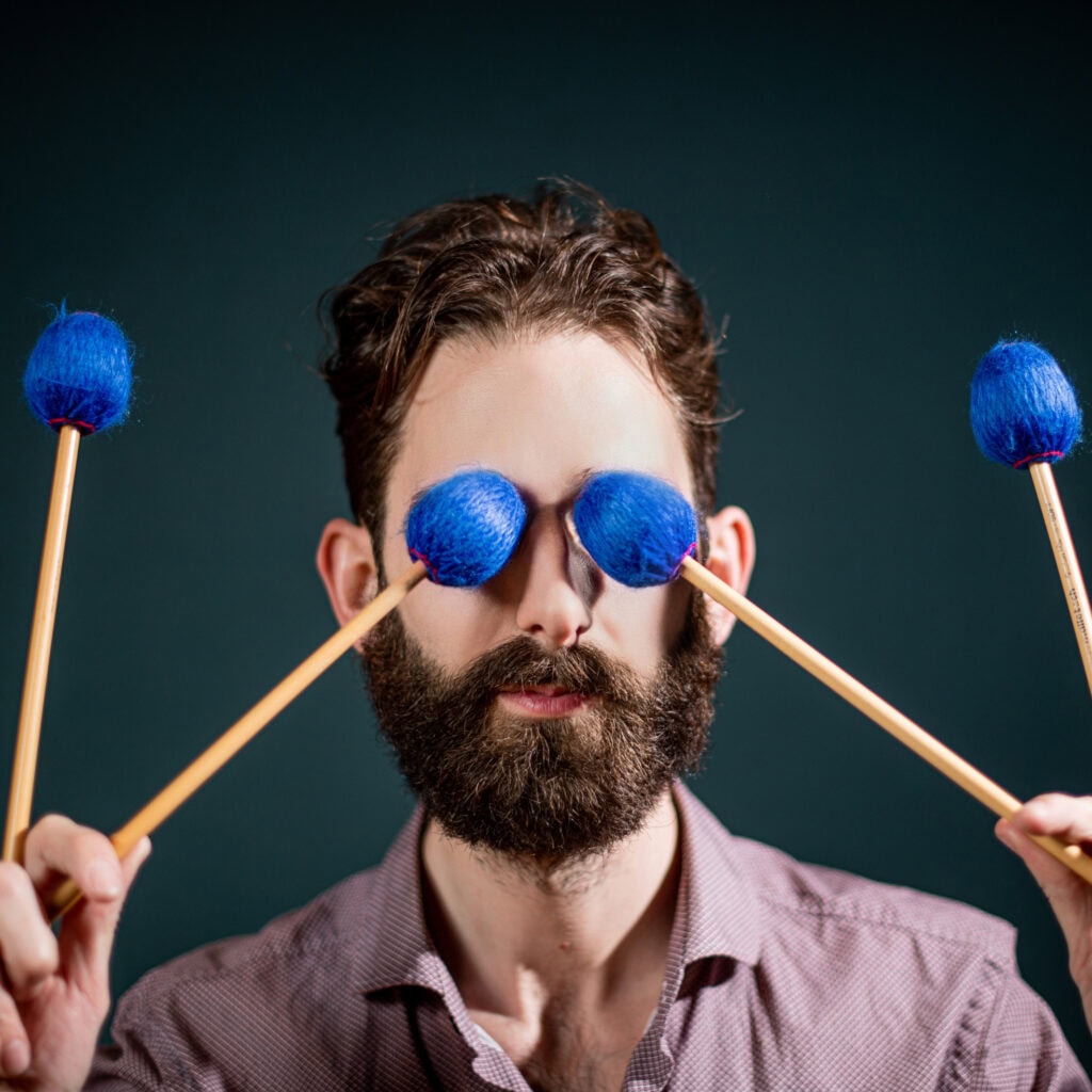 Farbenfrohes Portrait eines Mannes mit Schlagzeugstöcken, die druckvoll blaue Wollbälle an den Spitzen haben, kreativ inszeniert gegen dunklen Hintergrund.