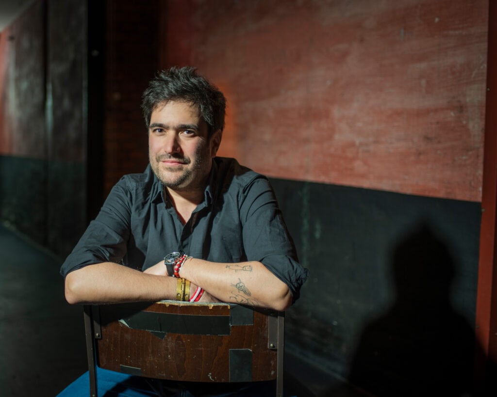 Mann mit dunklem Hemd sitzt an einem Tisch, lächelt in die Kamera, im Hintergrund eine rote Wand.