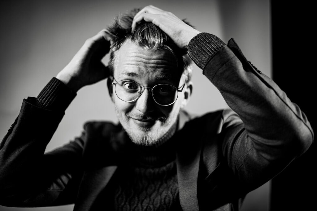 Lebensfrohe Porträtaufnahme eines jungen Mannes mit Brille, der sich die Haare stylt.