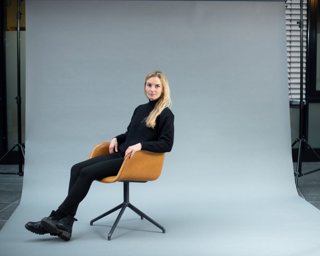 Hochwertiges Studiofoto einer jungen Frau in schwarz, sitzend auf einem modernen, braunen Drehstuhl vor grauem Hintergrund, professionell fotografiert von Frank Siemers aus Hamburg.