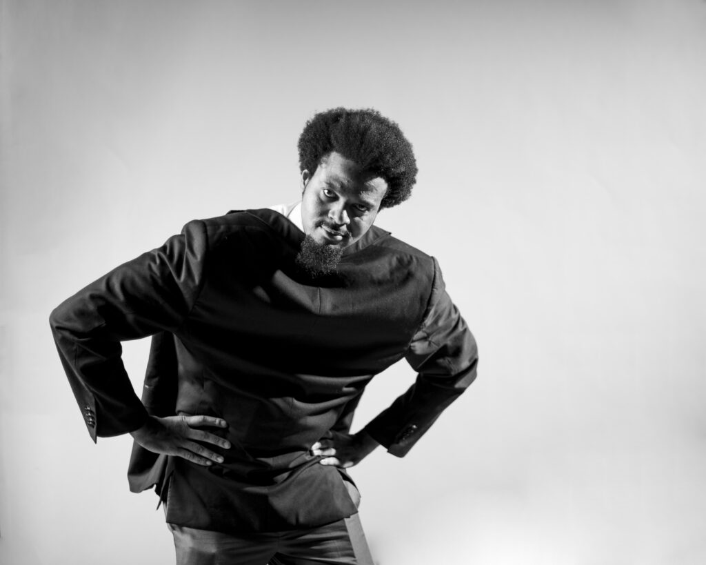 Stilvolle Schwarzweißaufnahme eines Mannes mit Afro-Frisur und Bart, der modisch gekleidet ist und in einer künstlerischen Pose auf einem blanken Hintergrund steht.