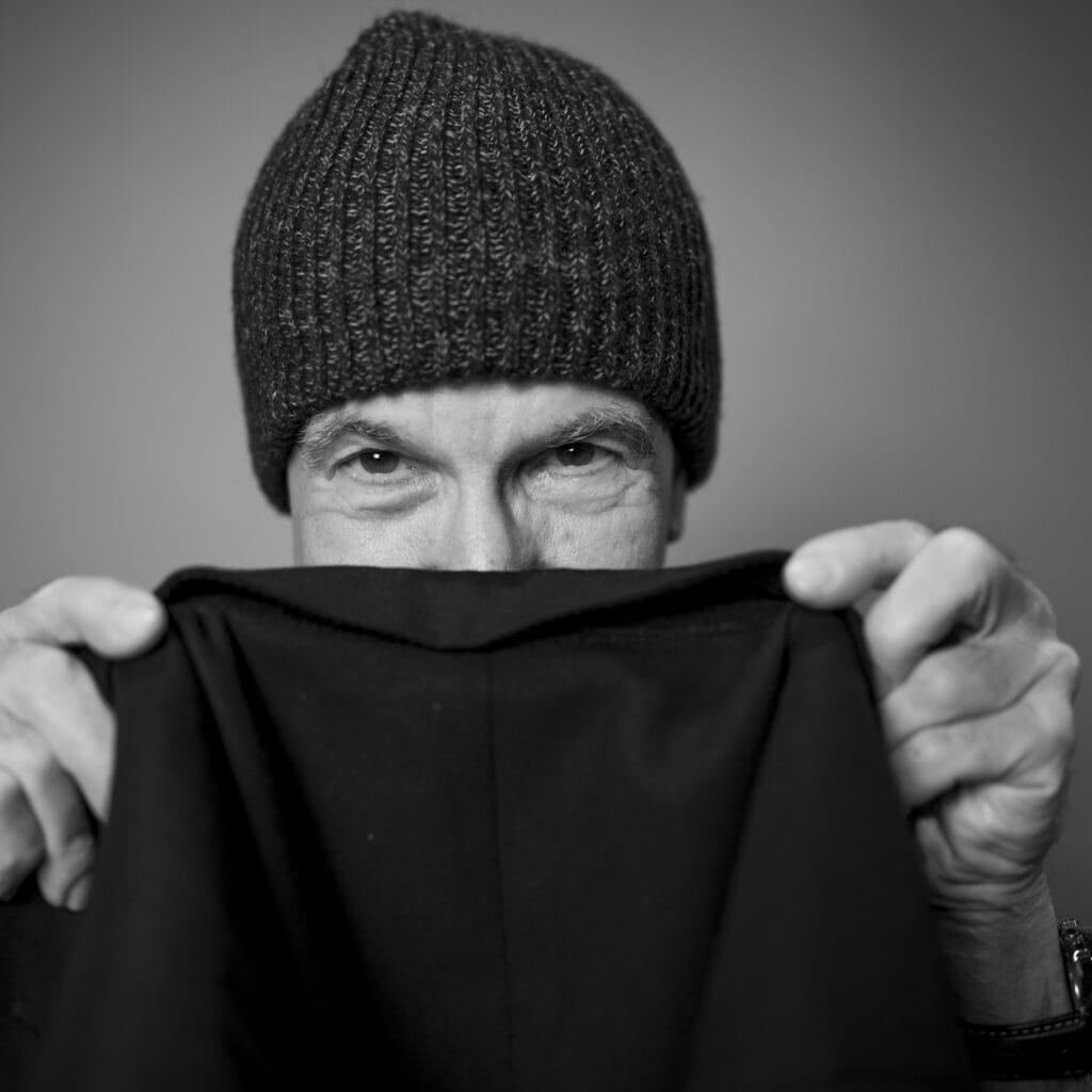 Ein Mann trägt eine dunkle Wollmütze und hält einen schwarzen Stoff vor seinem Gesicht, bei dem nur seine Augen sichtbar sind, in schwarz-weiß aufgenommen.