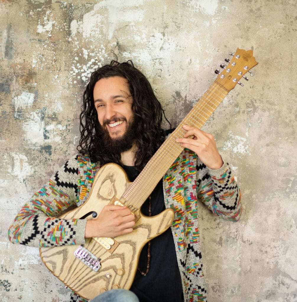 Frei schwingender Musiker mit langen Haaren und Bart, der eine selbstgebaute E-Gitarre vor einer Wand mit Rissen und Farbflächen hält.