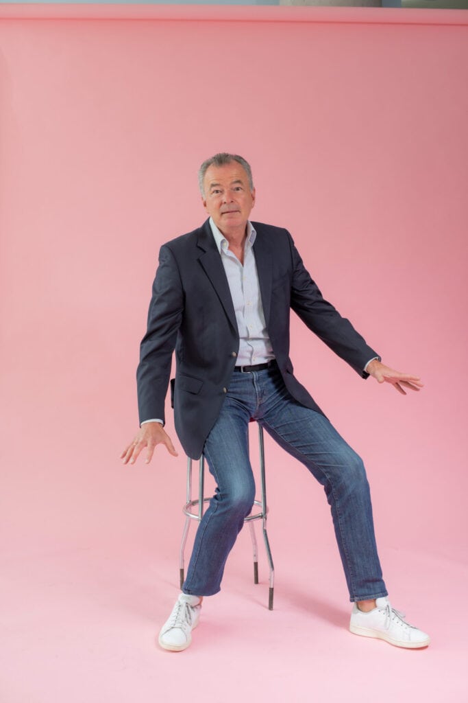 Frecher Mann im Anzug sitzt auf einem Hocker vor pinkem Hintergrund, trägt Jeans und Sneaker, wirkt entspannt und humorvoll in einem professionellen Fotoshooting von Frank Siemers.