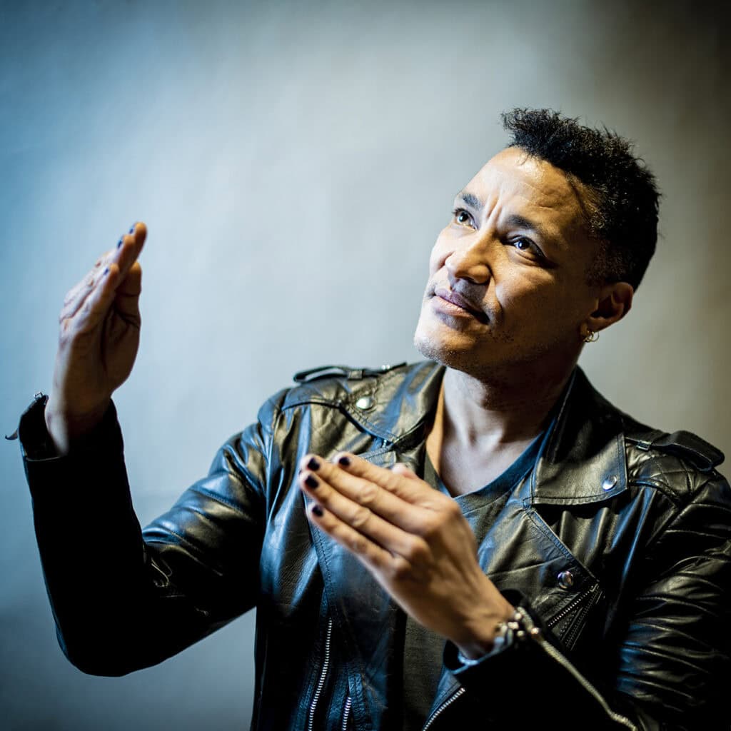 Künstlerporträt eines Mannes mit dunkler Haut, schwarzen Haaren und Lederjacke, der expressive Gesten macht und in die Kamera schaut.