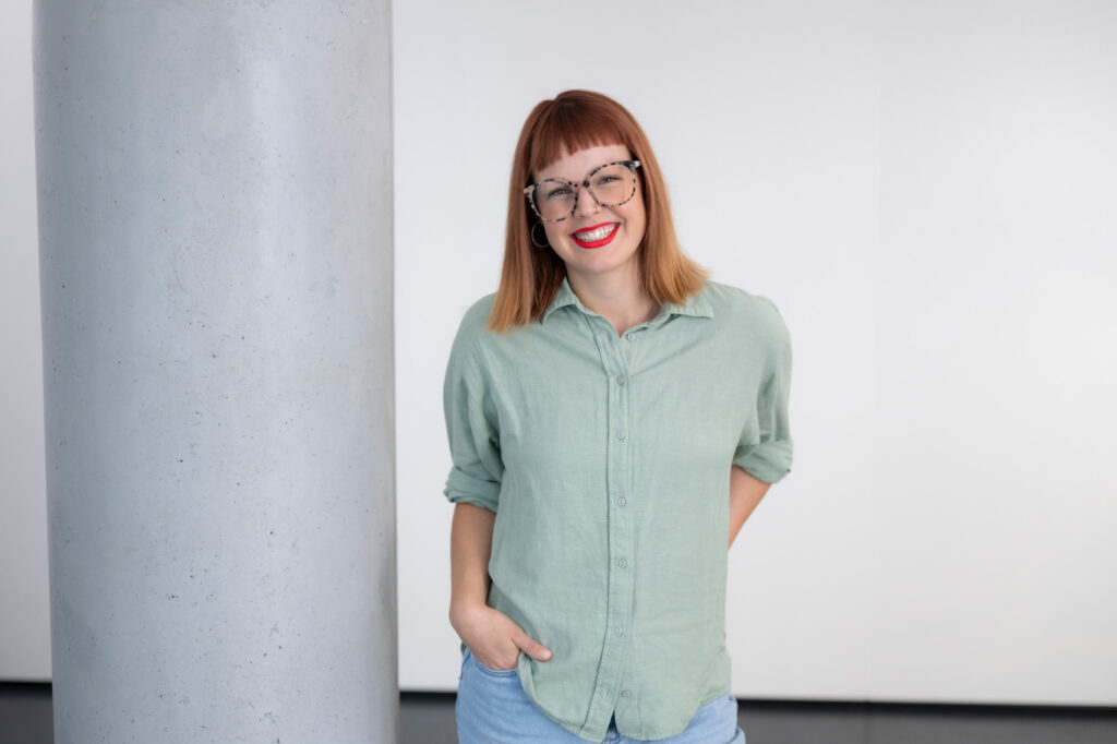 Authentische Porträtaufnahme einer jungen Frau mit roten Haaren, Brille und rotem Lippenstift, lächelnd vor einem weißen Hintergrund.