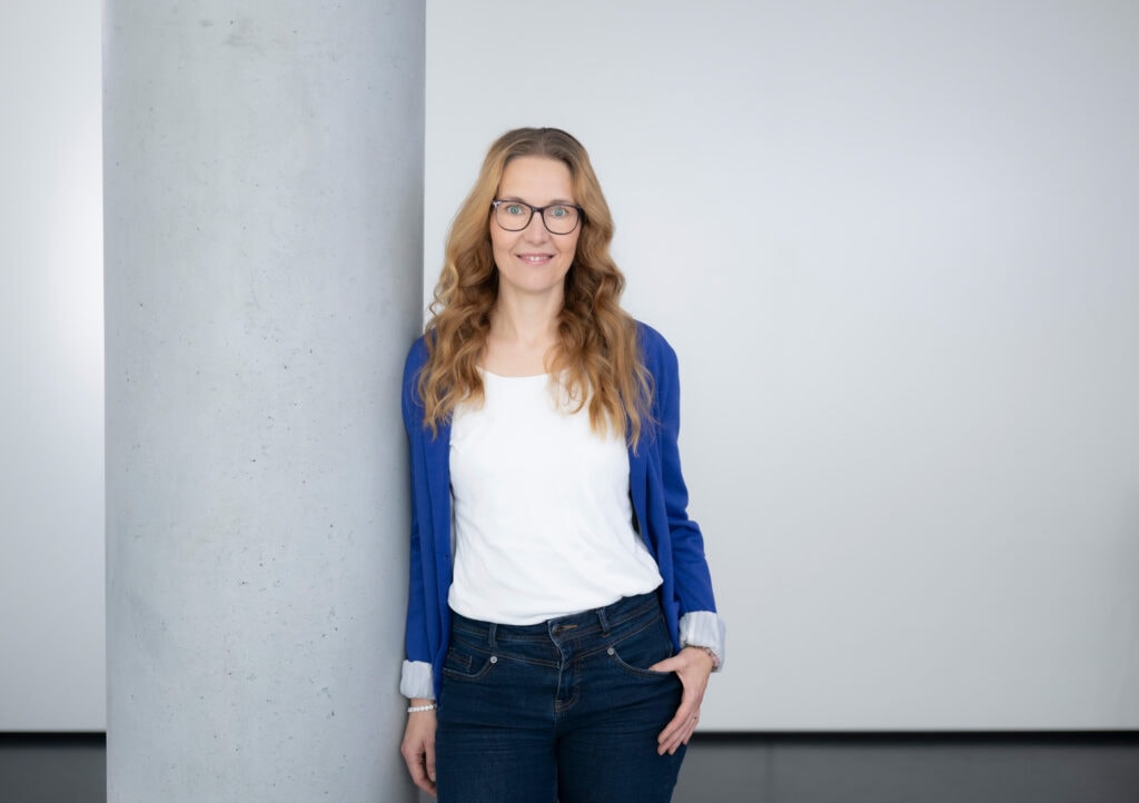 Hochwertiges Porträt eines Frau mit Brille, die neben einer Betonstütze steht, vor hellem Hintergrund, aufgenommen von Frank Siemers, professioneller Fotograf aus Hamburg.