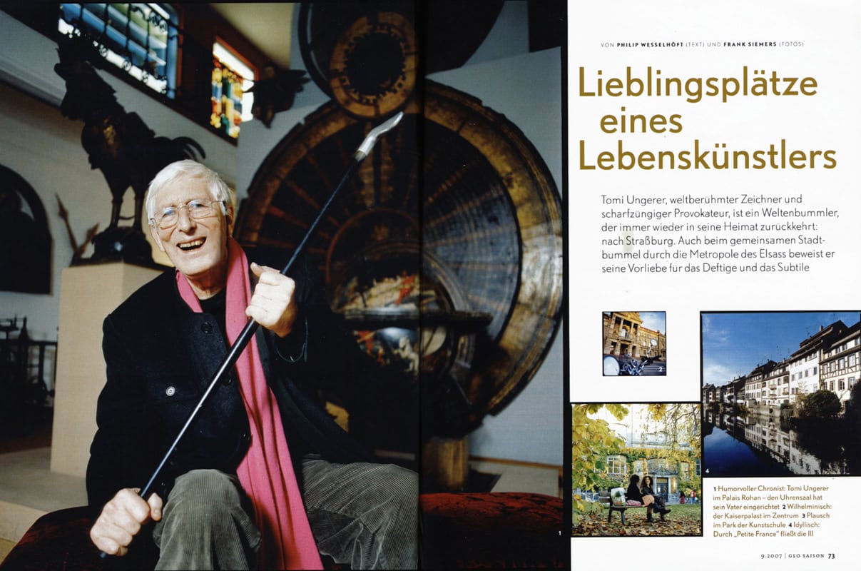 Tomi Ungerer, lachender älterer Mann mit Brille, hält Golfstock, im Hintergrund Kunstwerke und ein Museum.