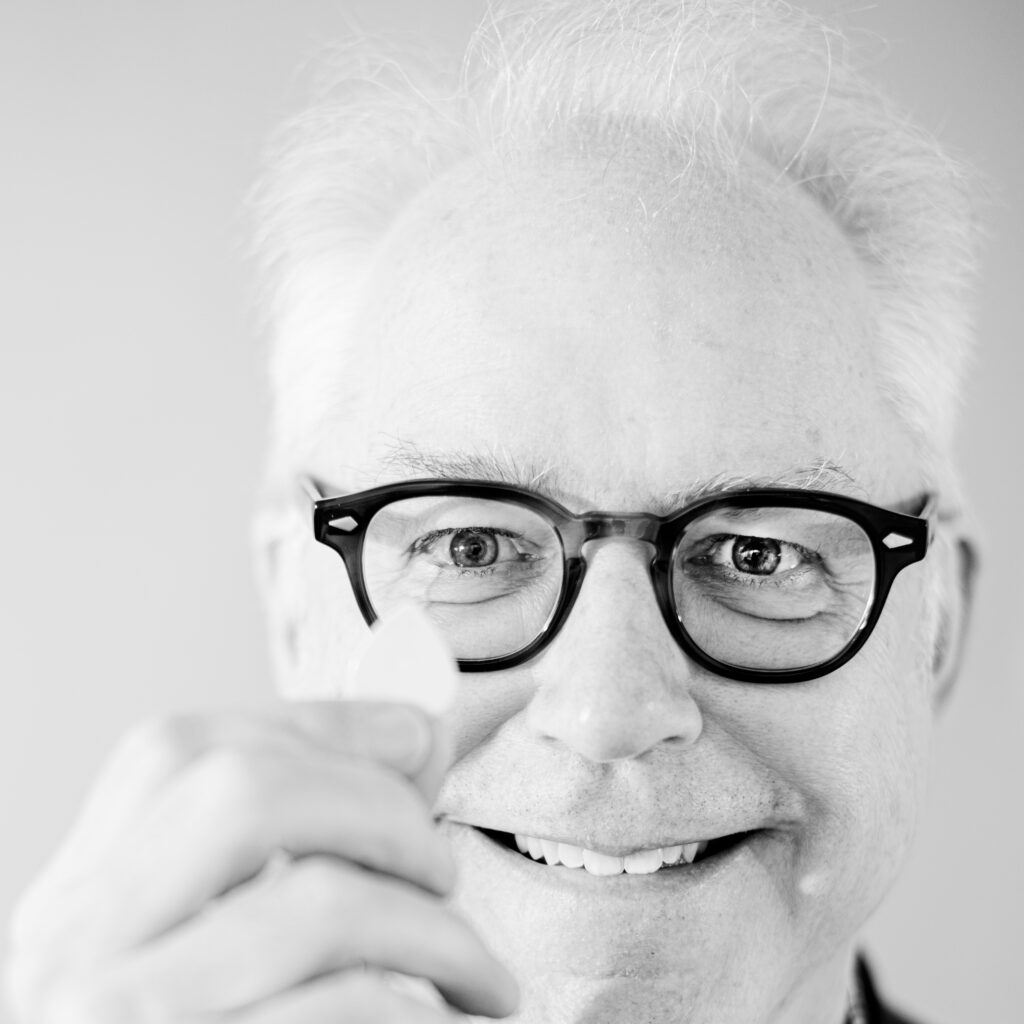 Hochwertiges Schwarz-Weiß-Porträt eines älteren Mannes mit schwarzen Brillengläsern und weißem Haar, der freundlich lächelt und direkt in die Kamera schaut.